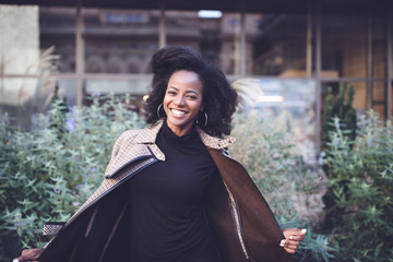 Beautiful african american young woman with afro and large hoop earrings in a stylish coat, smiling. Urban street portrait.