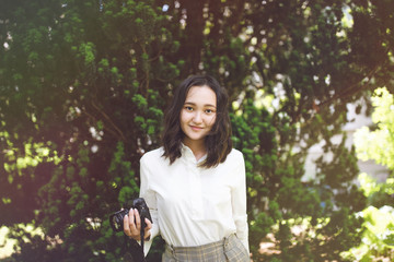 Optimistic asian girl in white shirt in a park, smiling, holding camera. Outdoor portrait.