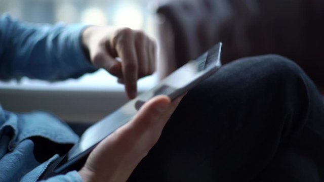 Close-up of a young business man using tablet sitting in a chair in a modern office opposite a window. Face of man is not shown. Tracking shot in slow motion.