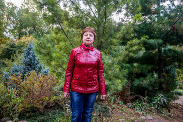 A lady in a red jacket and jeans stands on the background of coniferous trees