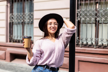 Young woman in black hat with coffee in hands walking in the street. Urban