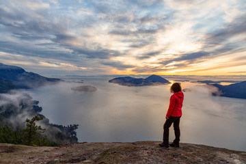 Adventurous Caucasian Girl sitting on top of a mountain during a colorful winter sunset. Taken on Tunnel Bluffs Hike, North of Vancouver, BC, Canada.