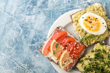 Three open sandwiches with avocado, red fish, egg and micro greenery on a white plate. Blue stone background. Close-up