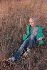 Happy smiling young blond woman with short haircut sitting on dry grass on lawn in the setting sun. Enjoy nature