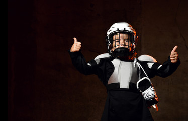 Positive boy in hockey uniform and protective helmet with hockey skates standing and showing thumb...