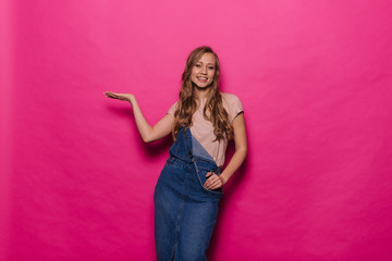 Smiling beautiful young woman holding and presenting copy space on her palm on isolated pink background. Your product presentation and advertising concept. Happy girl in casual denim clothes in studio