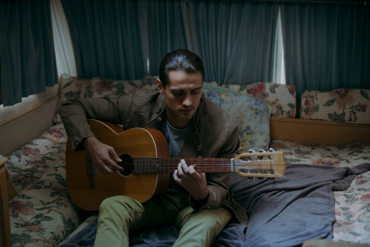 handsome hipster guy in neutral clothes sits on the bed and plays the guitar