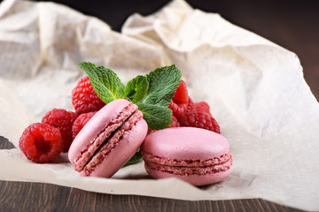 Pink macaroons with raspberries and mint leaves on a sheet of paper over a wooden background.