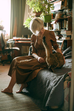 girl in a brown dress blonde strokes a cat on the couch at home
