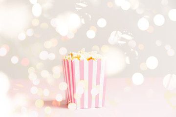 Popcorn bag with white and pink stripes at the festive lights bokeh backdrop. Empty minimalistic background. Romantic movie night and snack concept. Horizontal