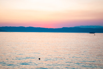 Sunset above Garda lake with floating ships, freshwater birds resting on water surface and dolomite mountains on the horizon. Lombardy, Italy