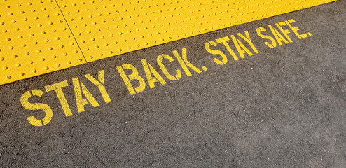diagonal view yellow sign at train station asking people to stay back, stay safe, on concrete floor
