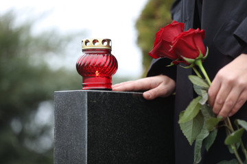Woman holding red roses near black granite tombstone with candle outdoors, closeup. Funeral ceremony