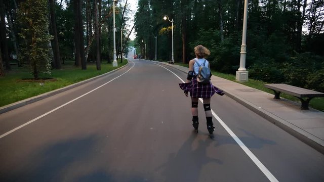A young curly blonde girl is quickly roller skating on a track in the park in the evening. The camera follows an active athletic woman on roller skates and in protective gear.