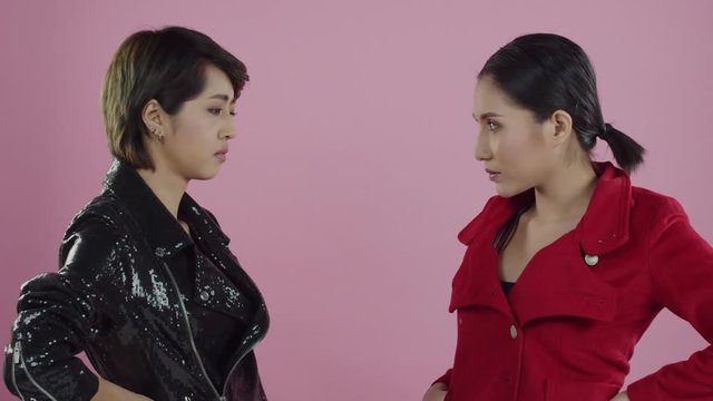 Young, attractive Asian women become angry and push each other with jealous and fierce expressions on thier faces - pink backdrop