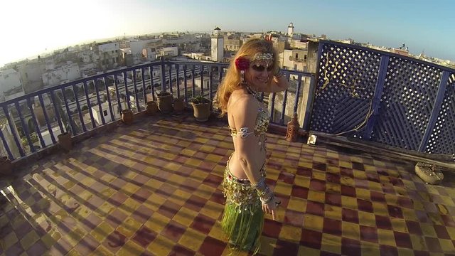 Camera orbits around beautiful belly dancer as she wiggles on the roof a building in Morocco at sunset.