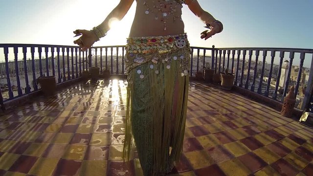 Backlit belly dancer doing hip thrusts on the roof of a building in Morocco at sunset.
