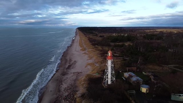 Aerial view of white painted steel lighthouse located in Pape, Latvia at Baltic sea coastline in cloudy day, wide angle establishing drone shot moving backwards