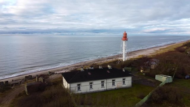 Aerial view of white painted steel lighthouse located in Pape, Latvia at Baltic sea coastline in cloudy day, wide angle establishing drone shot moving forward