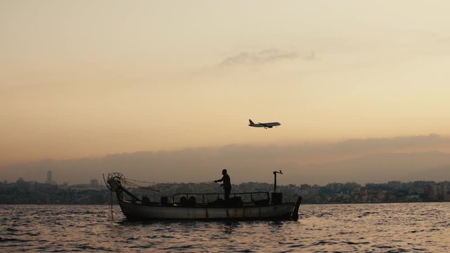 BEIRUT, LEBANON - CIRCA 2019: A Lebanese fisherman pulls a fishing net during sunrise. An airplane landing in the near airport. Beirut and Mount Lebanon in the horizon