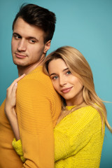 My real man. Close up photo of a young girl, who is standing in profile in a yellow outfit, hugging her boyfriend from the back, while he is trying to look at her.