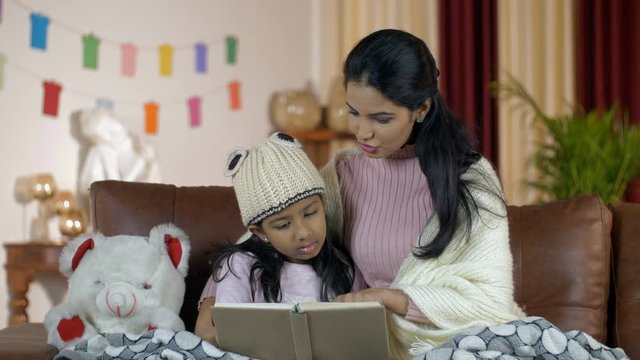 Beautiful Indian mother and daughter spending quality time together at home. Closeup shot of a young attractive mom reading a storybook to her cute little daughter during wintertime in India - fami...