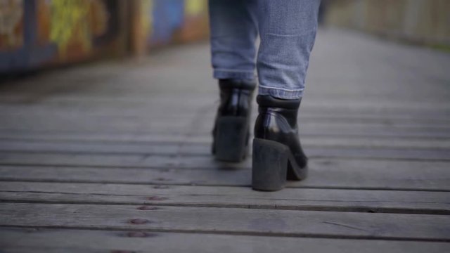 Close-up of women's legs in jeans and black heeled shoes, they walk on a wooden bridge, in the background a wall with graffiti.