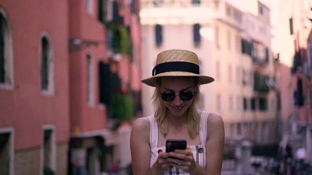Slow motion effect of cheerful hipster girl in stylish sunglasses typing text message for sending to friend during leisure time on Venice urban setting,  concept of technology and communication