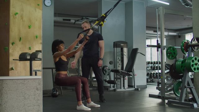 Determined sporty fit mixed race female exercising for endurance with trx fitness straps with personal trainer at gym. Beautiful woman training upper body on suspension trainer sling in health club.