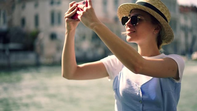 Cheerful female tourist in stylish sunglasses photographing on cellphone architect buildings during boat excursion around Venice city using mobile camera. Young caucasian woman traveler enjoying Italy