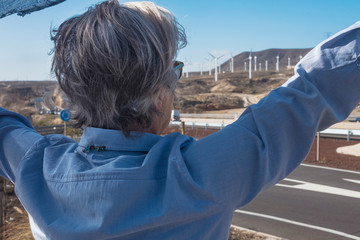 Fototapeta na wymiar Rear view of a senior woman with gray hair looking at the wind turbine plant in front to her. Mountain landscape. One caucasian people