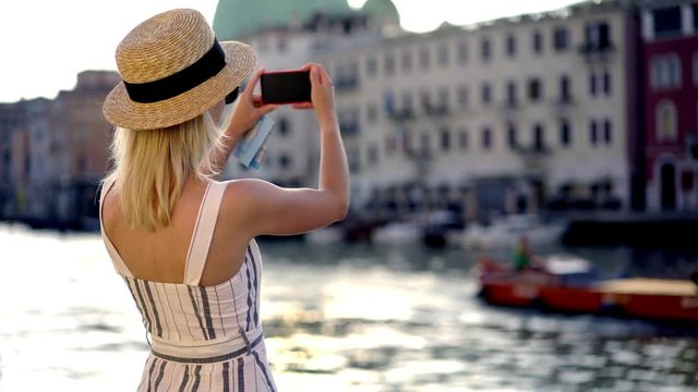 Caucasian female tourist in stylish hat making video for posting in travel web blog using smartphone camera during italian holidays, young hipster girl taking pictures of float boat on Venice canal ri