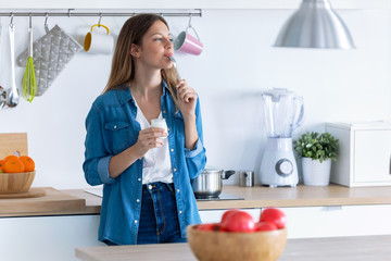 Pretty young woman eating yogurt while standing in the kitchen at home.