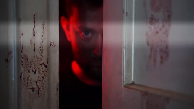 Crazy Horrible Serial Killer In Dark Looking in Door Gap. Imprint of a bloody hand on the door. Horror and red flashlights background