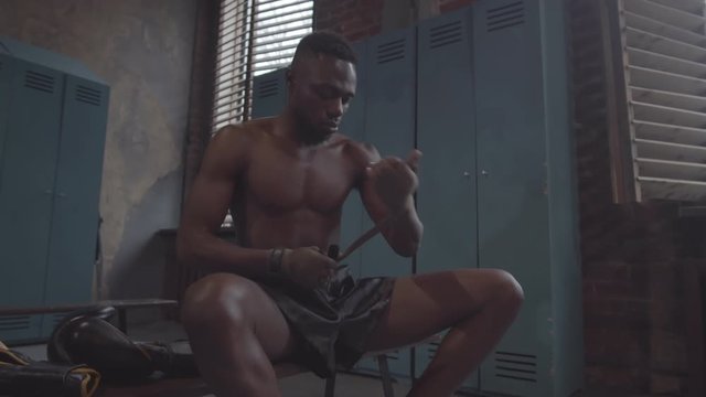 Knee-up shot of young African American boxer with bare chest, in black shorts sitting in dingy locker room at gym and wrapping wrists with handwraps, and boxing gloves lying on bench