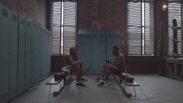 Full dollying shot of two athletic young African American fighters with bare chests sitting on bench in dingy gym changing room, getting ready for sparring, securing wrist wraps on hands and chatting