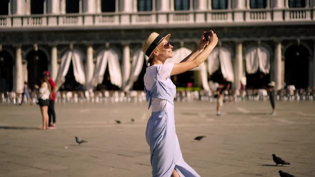 Slow Motion effect of happy smiling female traveller in summer dress exploring San Marco Square and taking pictures via application on modern cellular gadget for sharing photo in social networks