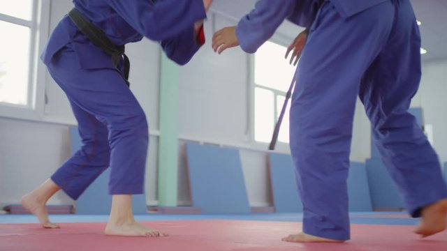 Young female jiu-jitsu athlete in blue sports uniform sitting on mat during break. Male partner walking in gym, helping her to stand up and start sparring fight
