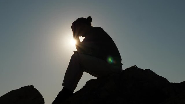 Tired or disappointed man silhouette, sit on stone against bright. He bend head down on hands, shake and put back again. Bad time (news) or problems concept