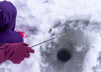 The hand of a gloved angler girl in winter holds a small fishing rod with a nod to a hole in the ice for the purpose of catching fish.