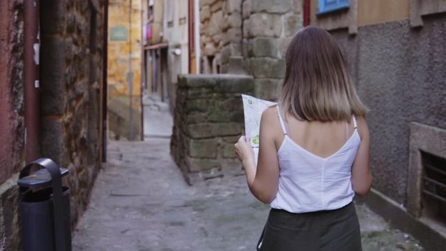 Young woman tourist is walking in old narrow street in Porto, Portugal, admiring ancient architecture, searching landmarks and intersting spots with the help of paper map, view from the back.