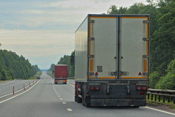 Big Semi trucks with white semi-trailer driving on suburban empty highway road on Summer day on green trees and cloudy sky background, back view