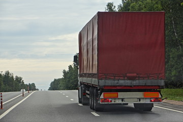 Big Semi truck with maroon van driving on suburban empty highway road on Summer day on green trees...