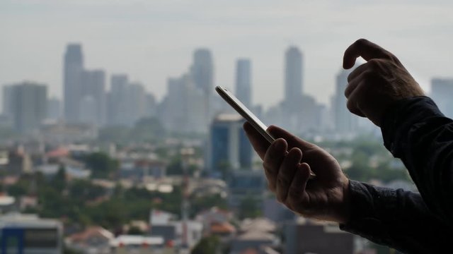 A man dials a message on a smartphone against the background of the panorama of the city of Jakarta. Hands close up.
