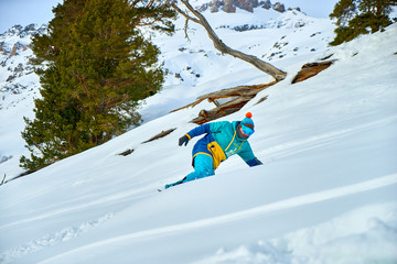 A young man in a colored winter hoodie and mask rides in the mountains on a snowboard in a cool hat with an orange pom pom