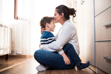 Mom and her son hugging each other. Mom is kissing her kid in the forehead. - obrazy, fototapety, plakaty