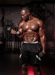 Fototapeta na wymiar Muscular African American shirtless male bodybuilding athlete does dumbbell curls in a dark grungy gym with dramatic lighting flare 