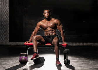 Muscular African American shirtless, sweaty male bodybuilding athlete sitting on a bench showing his six pack abs  in a dark grungy gym with dramatic lighting flare 