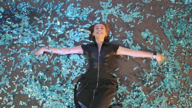 Crazy New Year's Eve. Slow motion A beautiful young woman lies on the floor and creates a snow angel out of blue confetti