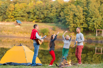 Group of happy friends with guitar, having fun outdoor, dancing and jumping near the lake in the park background the Beautiful sky. Camping fun